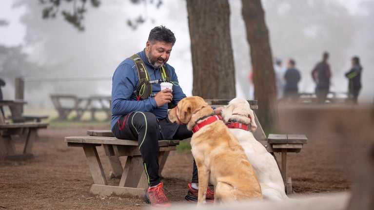 Man sitting outside with two dogs, holding a takeaway cup of coffee